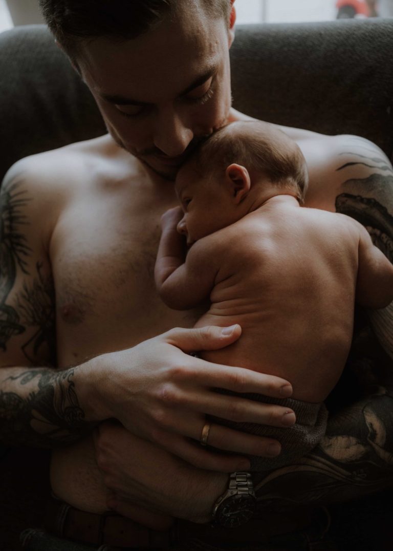 Ein tätowierter Vater hält sein schlafendes Baby auf der Brust.